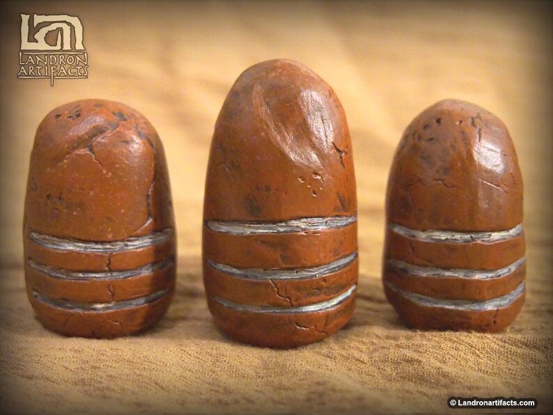 May include: Three brown, egg-shaped artifacts with horizontal silver bands. The objects have a textured surface and a weathered appearance. The artifacts are displayed on a tan surface. The Landron Artifacts logo is in the upper left corner.