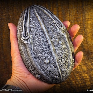 May include: A detailed, silver-toned, oval-shaped artifact with intricate patterns and textures. The object is held in a hand, showcasing its size and design. The surface features raised, organic-looking details. The Etsy shop name, Landron Artifacts, is visible.