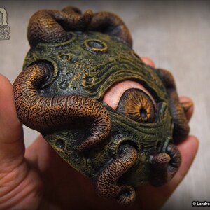 May include: A green and brown, textured, eye-like sculpture with a single large eye and multiple tentacles. The sculpture is made of resin and is approximately 4 inches in diameter.