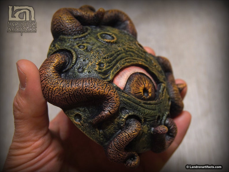 May include: A green and brown, textured, eye-like sculpture with a single large eye and multiple tentacles. The sculpture is made of resin and is approximately 4 inches in diameter.