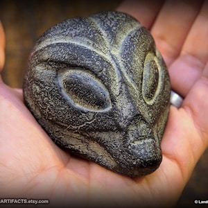 May include: A dark gray alien head sculpture with large oval eyes and a textured surface. The sculpture is held in a hand, showcasing the intricate details of the alien's features. The Etsy shop name is visible.