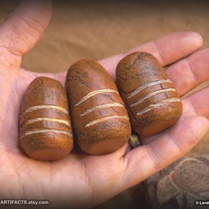 May include: Three brown, oblong-shaped stones with horizontal white stripes are displayed in a hand. The stones vary slightly in size and have a textured surface. The Etsy shop name, LANDRON ARTIFACTS, is visible in the image.