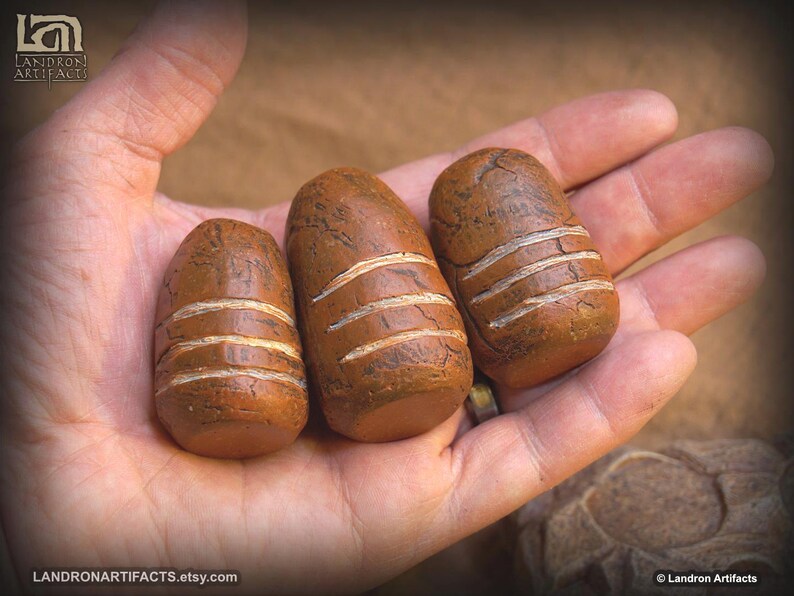 May include: Three brown, oblong-shaped stones with horizontal white stripes are displayed in a hand. The stones vary slightly in size and have a textured surface. The Etsy shop name, LANDRON ARTIFACTS, is visible in the image.