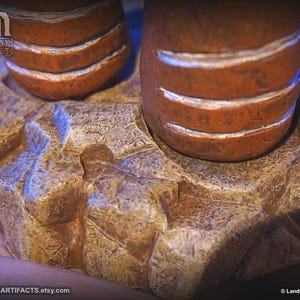 May include: Close-up of a handcrafted, textured sculpture. The sculpture features two cylindrical, orange-brown forms with white bands, resting on a detailed, rock-like base. The base is a mix of tan and gold tones.