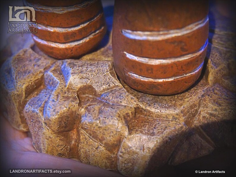 May include: Close-up of a handcrafted, textured sculpture. The sculpture features two cylindrical, orange-brown forms with white bands, resting on a detailed, rock-like base. The base is a mix of tan and gold tones.