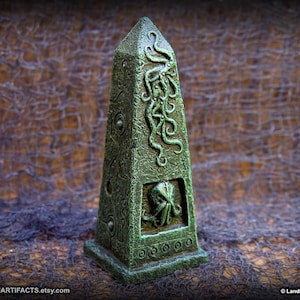 May include: A green obelisk-shaped sculpture with intricate carvings of tentacles and a creature's face. The sculpture has a textured surface and is set against a dark background. The base of the obelisk has a series of small, circular designs.