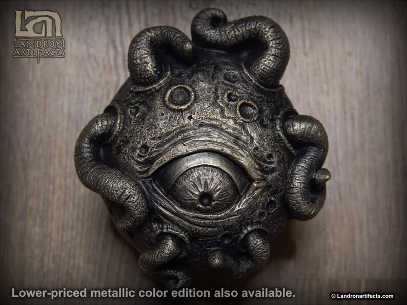 May include: A bronze-colored, detailed, three-dimensional sculpture of a single eye with tentacles. The eye is large and prominent, with a detailed iris and pupil. The tentacles are curled and twisted, giving the sculpture a menacing appearance.