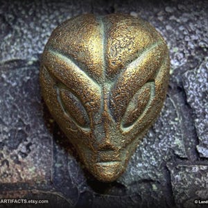 May include: A bronze-colored alien head sculpture with detailed facial features, including large almond-shaped eyes and a defined chin. The sculpture has a textured surface and is set against a dark, stone-like background. The Etsy shop name is visible.