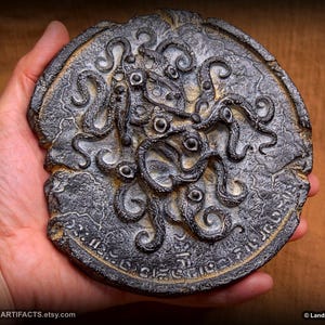 May include: A dark, weathered, circular artifact with an intricate design of tentacles and eyes. The surface has a textured, aged appearance with etched details. The artifact is held in a hand, showcasing its size and craftsmanship. The text "LANDRON ARTIFACTS" is visible.