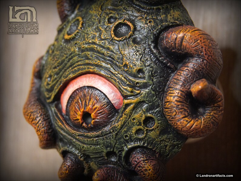 May include: A green and brown, textured, sculpted, one-eyed creature with a large, round, orange eye. The creature has three large, curled, brown tentacles.