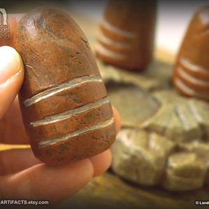 May include: A hand holds a brown, cylindrical artifact with three horizontal, white carved lines. The artifact has a weathered appearance. The background shows similar objects and a carved stone base. The Etsy shop name is visible.