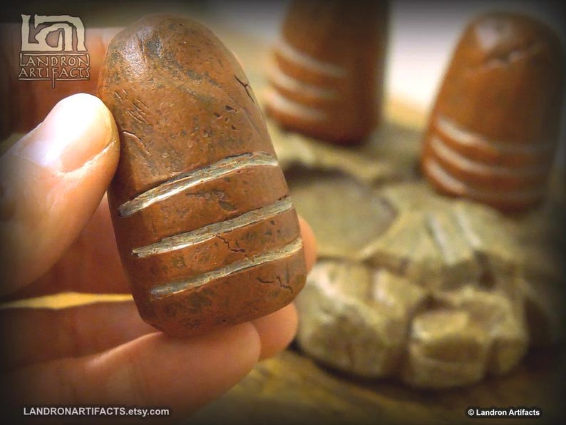 May include: A hand holds a brown, cylindrical artifact with three horizontal, white carved lines. The artifact has a weathered appearance. The background shows similar objects and a carved stone base. The Etsy shop name is visible.