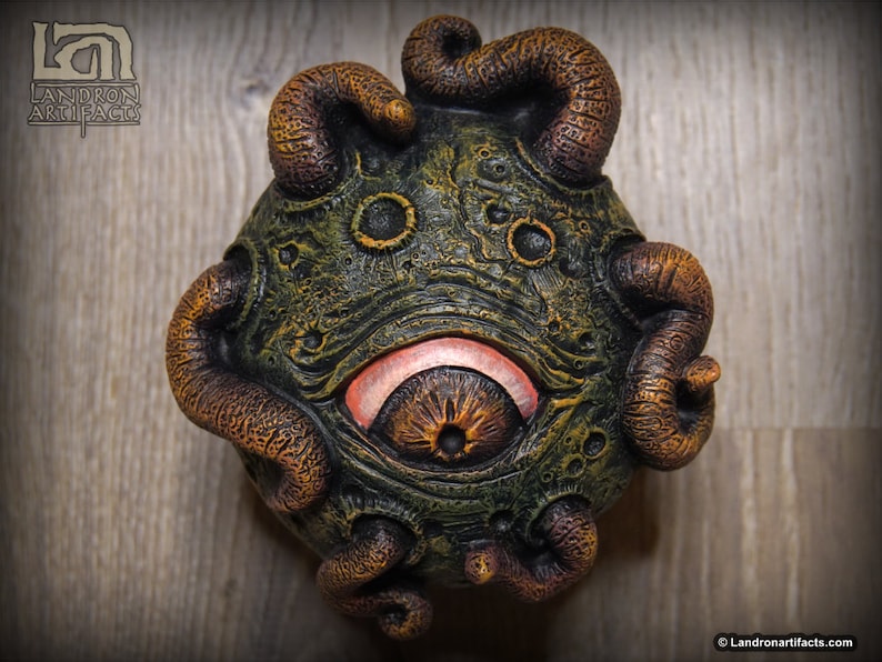 May include: A green and brown, textured, spherical sculpture with a large, orange eye and several tentacles. The sculpture has a slightly menacing expression.