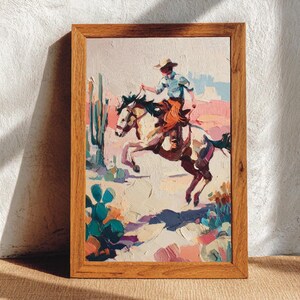 May include: A framed painting shows a cowboy on horseback in a desert scene. The artwork uses pink, blue, and green colours, highlighting the rider and horse. The wooden frame surrounds the painting.