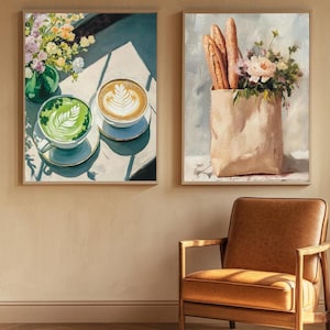 May include: Two framed paintings, one depicting a green and brown latte with a latte art design and the other depicting a brown paper bag with baguettes and flowers. The paintings are hanging on a beige wall above a brown leather armchair.