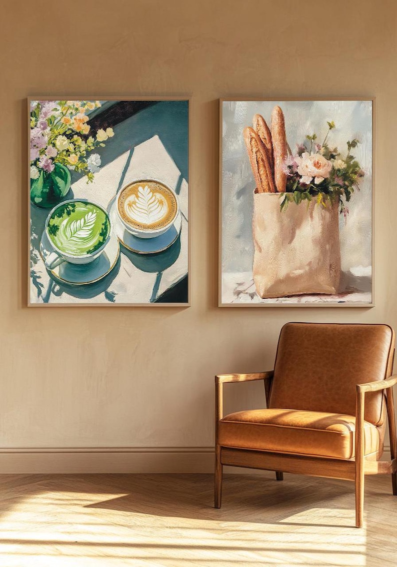 May include: Two framed paintings, one depicting a green and brown latte with a latte art design and the other depicting a brown paper bag with baguettes and flowers. The paintings are hanging on a beige wall above a brown leather armchair.