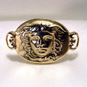 May include: A gold-toned brooch featuring a detailed relief of a classical female face within an oval frame. The brooch has ornate, curved accents on each side, and is likely made of metal.