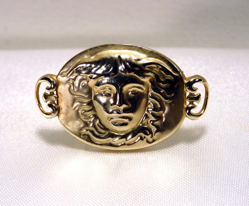 May include: A gold-toned brooch featuring a detailed relief of a classical female face within an oval frame. The brooch has ornate, curved accents on each side, and is likely made of metal.
