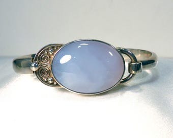 May include: A silver bracelet featuring a large, oval, pale blue gemstone. The bracelet has a decorative clasp with spiral and dot details. The gemstone is set in a silver bezel.