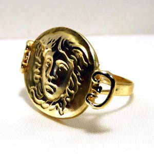 May include: A gold-colored ring featuring a detailed relief of a classical face within a circular setting. The ring has a band and decorative side elements, all in a polished gold tone. The design evokes ancient Greek or Roman aesthetics.