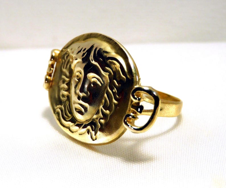May include: A gold-colored ring featuring a detailed relief of a classical face within a circular setting. The ring has a band and decorative side elements, all in a polished gold tone. The design evokes ancient Greek or Roman aesthetics.
