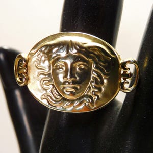 May include: A gold-toned ring featuring a detailed relief of a face within an oval frame. The ring has decorative side accents and is displayed on a black surface. The face has detailed features and flowing hair.