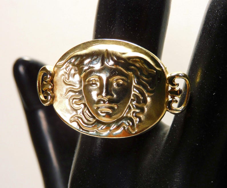May include: A gold-toned ring featuring a detailed relief of a face within an oval frame. The ring has decorative side accents and is displayed on a black surface. The face has detailed features and flowing hair.