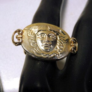 May include: Gold-toned ring featuring an oval medallion with a sculpted face. The ring has a detailed design with a classic aesthetic. The ring is worn on a finger.