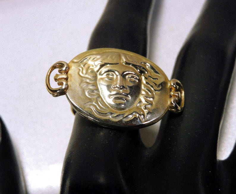 May include: Gold-toned ring featuring an oval medallion with a sculpted face. The ring has a detailed design with a classic aesthetic. The ring is worn on a finger.