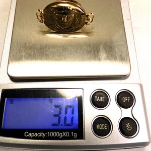 May include: A gold-colored ring with a detailed face design, resting on a digital scale. The scale displays a weight of 3.0 grams. The ring has an oval shape with decorative side elements.