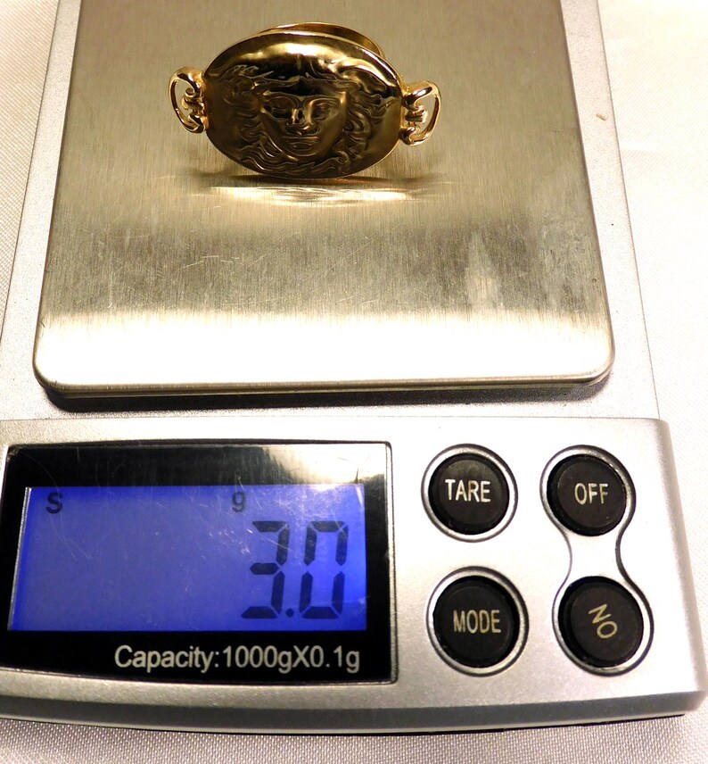May include: A gold-colored ring with a detailed face design, resting on a digital scale. The scale displays a weight of 3.0 grams. The ring has an oval shape with decorative side elements.