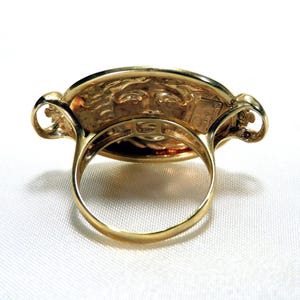 May include: A gold-colored ring with an oval face featuring a detailed relief of a face. The ring has a thick band and decorative elements on the sides of the face. The ring is set against a white background.