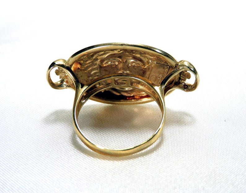 May include: A gold-colored ring with an oval face featuring a detailed relief of a face. The ring has a thick band and decorative elements on the sides of the face. The ring is set against a white background.