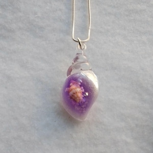 May include: A silver chain necklace with a purple and clear resin seashell pendant. The pendant contains a smaller seashell inside.