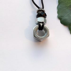 May include: A black cord necklace with a silver metal nut pendant and a black bead.
