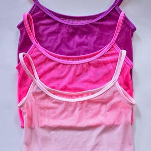 Puede incluir: Tres camisetas sin mangas transparentes en tonos rosa y morado apiladas. La camiseta superior es de color morado oscuro, la del medio es rosa brillante y la inferior es rosa claro. Cada una tiene tirantes finos y un escote redondo. Fondo blanco.