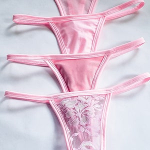 May include: Four pink thong underwear. The top three are solid pink, while the bottom one features a floral lace design on the front. The thongs are arranged vertically against a white background.