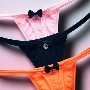 May include: Three thong underwear in pink, black, and orange. Each thong features a small black bow at the front with a decorative charm. The underwear is made of a smooth, possibly silky material, and is displayed against a pink background.