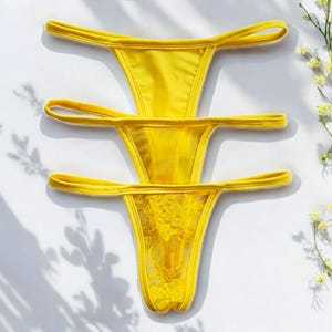 May include: Three yellow thong underwear arranged on a white surface. The top two are solid, while the bottom one features a lace panel. The image is lit with natural light, and the shadows of leaves and flowers are visible.