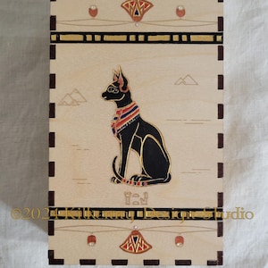 Peut inclure: Une boîte en bois avec un motif de chat noir. Le chat est assis, la tête tournée vers la gauche. La boîte est ornée d'une bordure dorée et d'un motif noir et or sur le dessus et le dessous. Le texte "2022 Kilkenny Design Studio" est inscrit sur le dessous de la boîte.