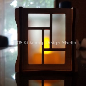 May include: A white wooden candle holder with a frosted glass insert and a geometric design. The candle holder is lit and casting a warm glow.
