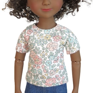 May include: A doll with dark skin, curly brown hair, and pink eyes. The doll wears a short-sleeved shirt with a floral pattern in pastel colors and blue denim pants. The doll's arms and legs are articulated.