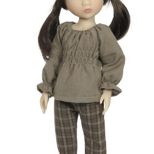 May include: A doll wearing a two-piece outfit. The top is a long-sleeved, taupe-colored blouse with a gathered waist and ruffled cuffs. The pants are brown plaid. The doll has brown hair in pigtails.