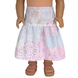 May include: A knee-length skirt featuring a patchwork design with various floral, gingham, and butterfly patterns in shades of light pink, lavender, and white. The skirt has a gathered waist and a flared hem, paired with light pink strappy sandals.