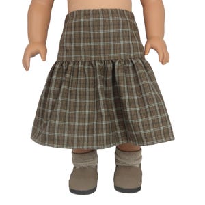 May include: A doll wearing a brown plaid skirt with a ruffled hem. The skirt has a brown, white, and light blue plaid pattern. The doll is also wearing matching brown shoes and socks.