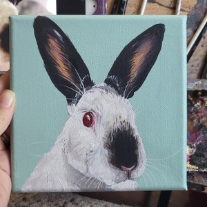 May include: A painting of a white rabbit with black ears and a black nose. The rabbit has red eyes and is looking to the right. The background is a light blue color.
