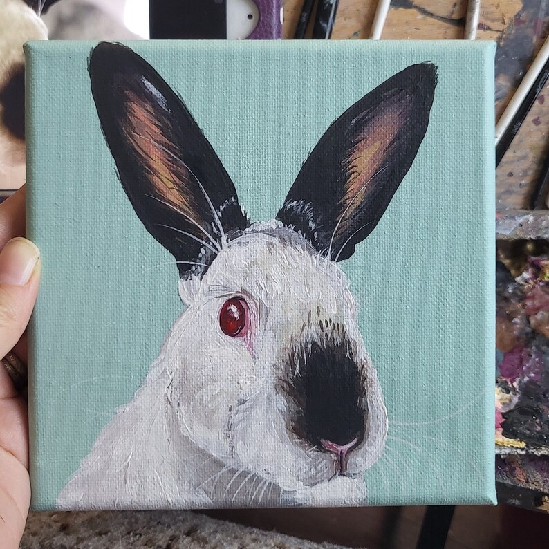 May include: A painting of a white rabbit with black ears and a black nose. The rabbit has red eyes and is looking to the right. The background is a light blue color.