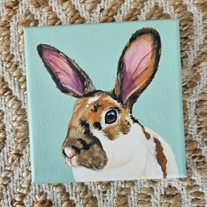 May include: A painting of a brown and white rabbit with pink ears on a light blue background.