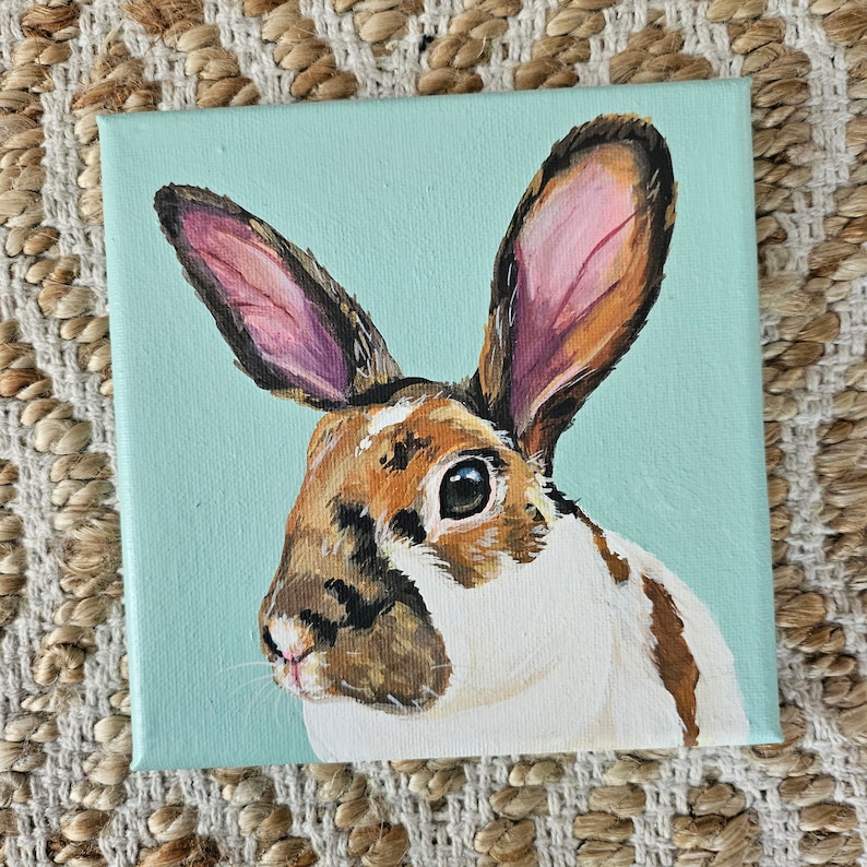 May include: A painting of a brown and white rabbit with pink ears on a light blue background.
