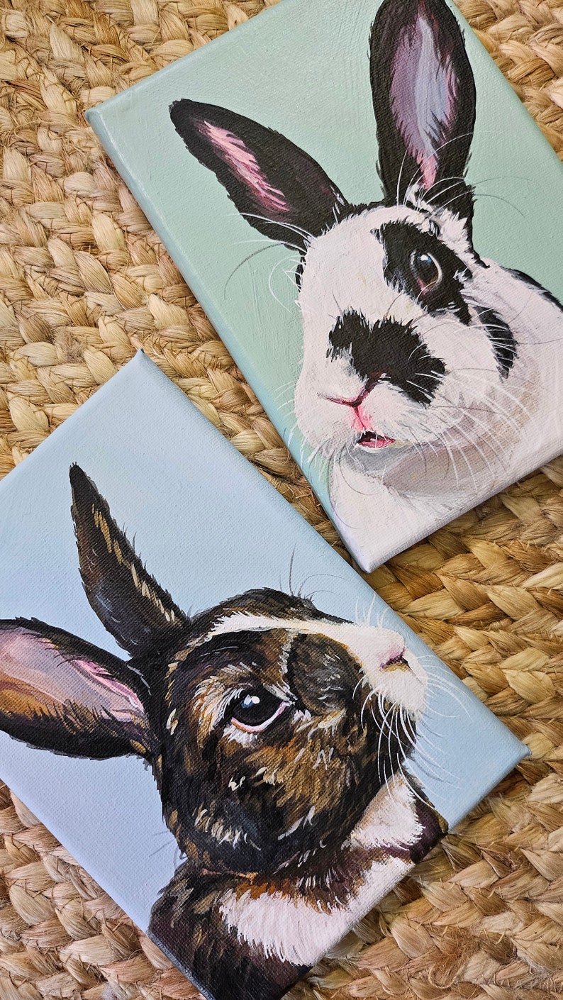 May include: Two paintings of rabbits on canvas. The first painting is of a black and white rabbit with pink ears on a light blue background. The second painting is of a brown and white rabbit with brown ears on a light blue background.